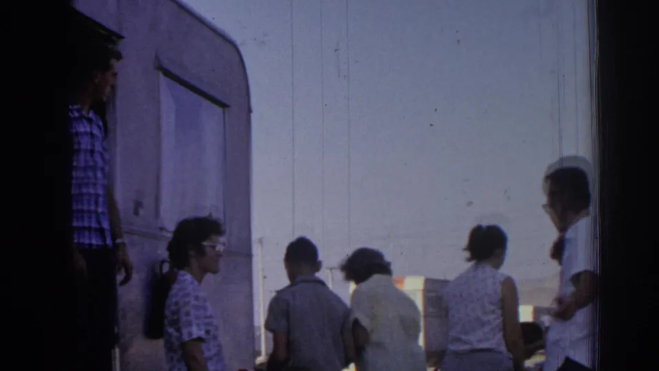 1962 Izmir campground frame with adults gathered beside a camper trailer, viewed in handheld 8mm footage.