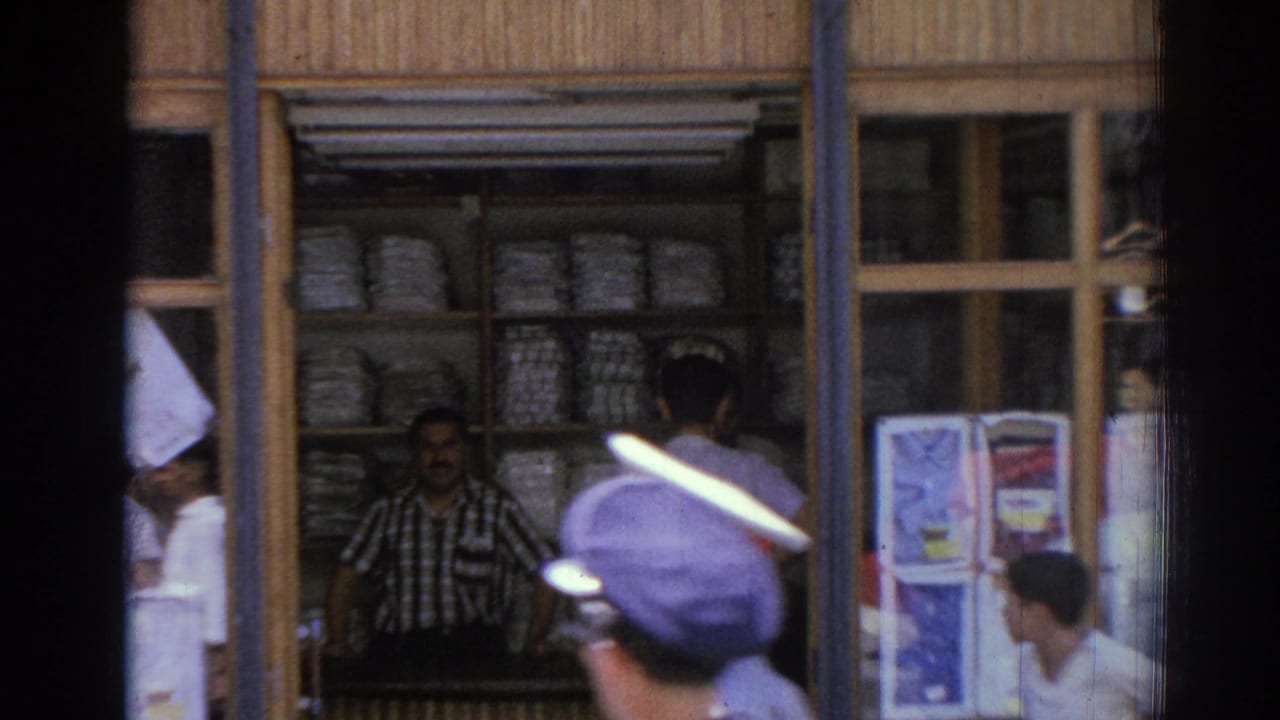 Street-side shop seen through glass with merchant behind stacked goods and a passerby in a peaked cap.