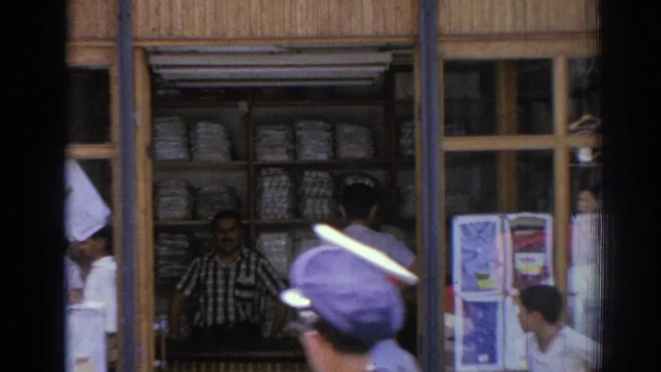 1962 Izmir street shop viewed through glass, with a checkered-shirt merchant, shelves of wrapped goods, and a peaked cap passerby in front.