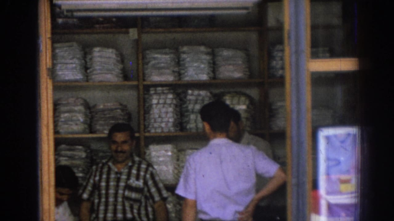Inside a 1962 Izmir shop, a merchant stands before shelves of bundled goods as a customer in a light shirt browses or speaks with him.