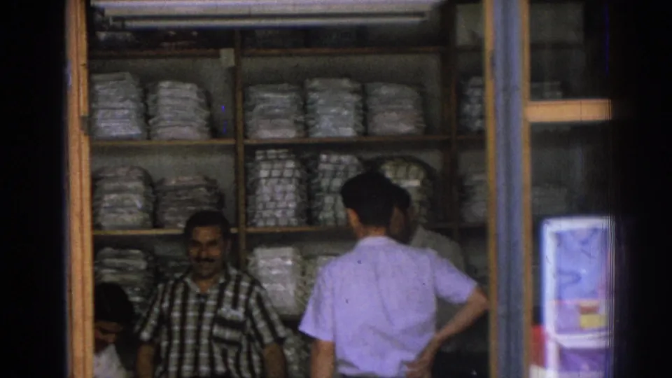 Inside a 1962 Izmir shop, a merchant stands before shelves of bundled goods as a customer in a light shirt browses or speaks with him.