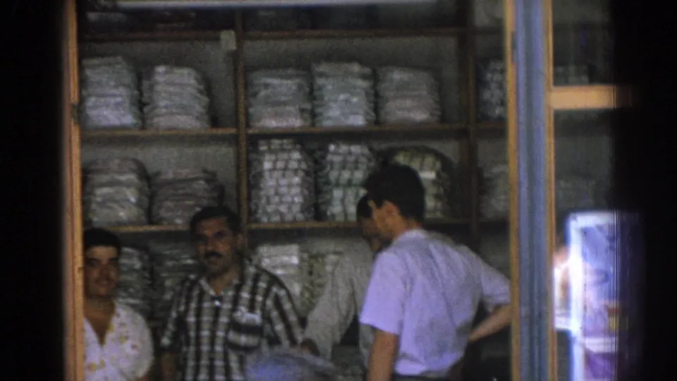 A 1962 Izmir storefront view shows a light-shirt customer beside a merchant, with shelves of wrapped goods filling the shop background.