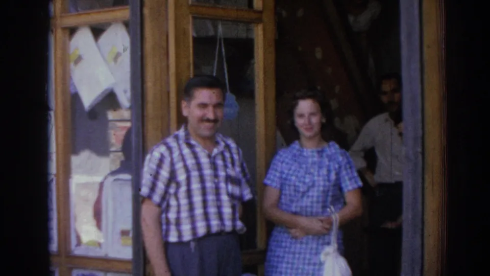 The home-movie camera catches the same smiling pair as another adult stands in the dim shop entrance.