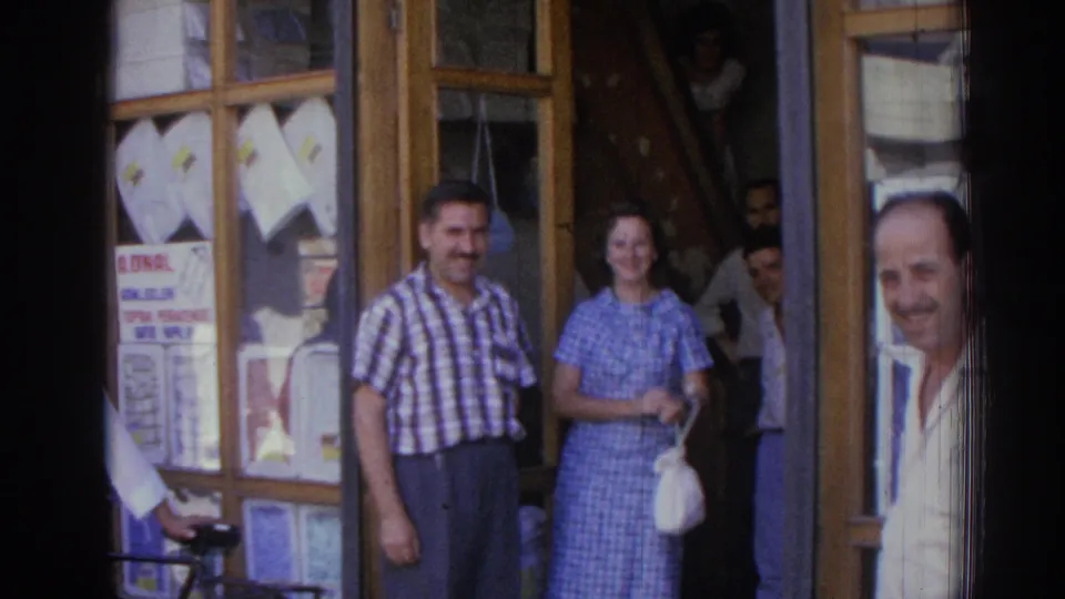 A wider 1962 Izmir street view shows the smiling pair, shop window, background men, and an older man entering at right.