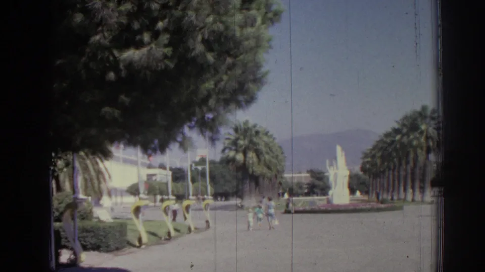 A wide 1960s Izmir park view shows palm-lined paths, a central fountain, trees, and pedestrians in bright daylight.