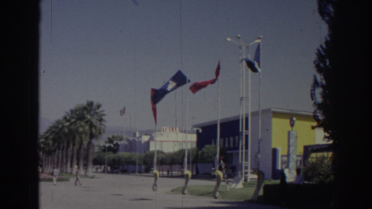 Flags fly beside a yellow-and-blue building with palm trees and pedestrians nearby.