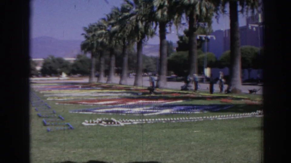 1962 Izmir home-movie frame showing colorful flower-bed patterns, tall palms, distant hills, and people walking in the park.