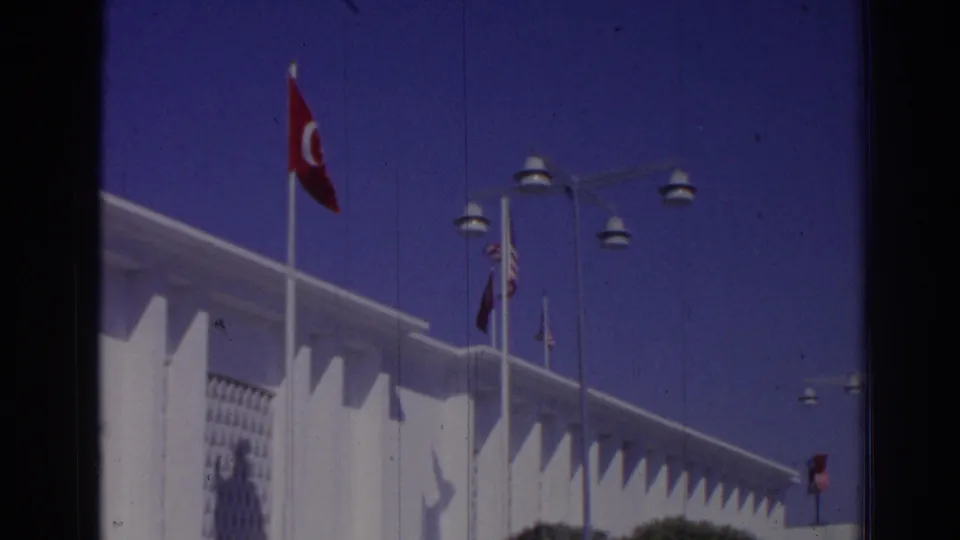 1962 Izmir home-movie frame showing a Turkish flag above a white modernist building with street lamps and geometric facade detail.
