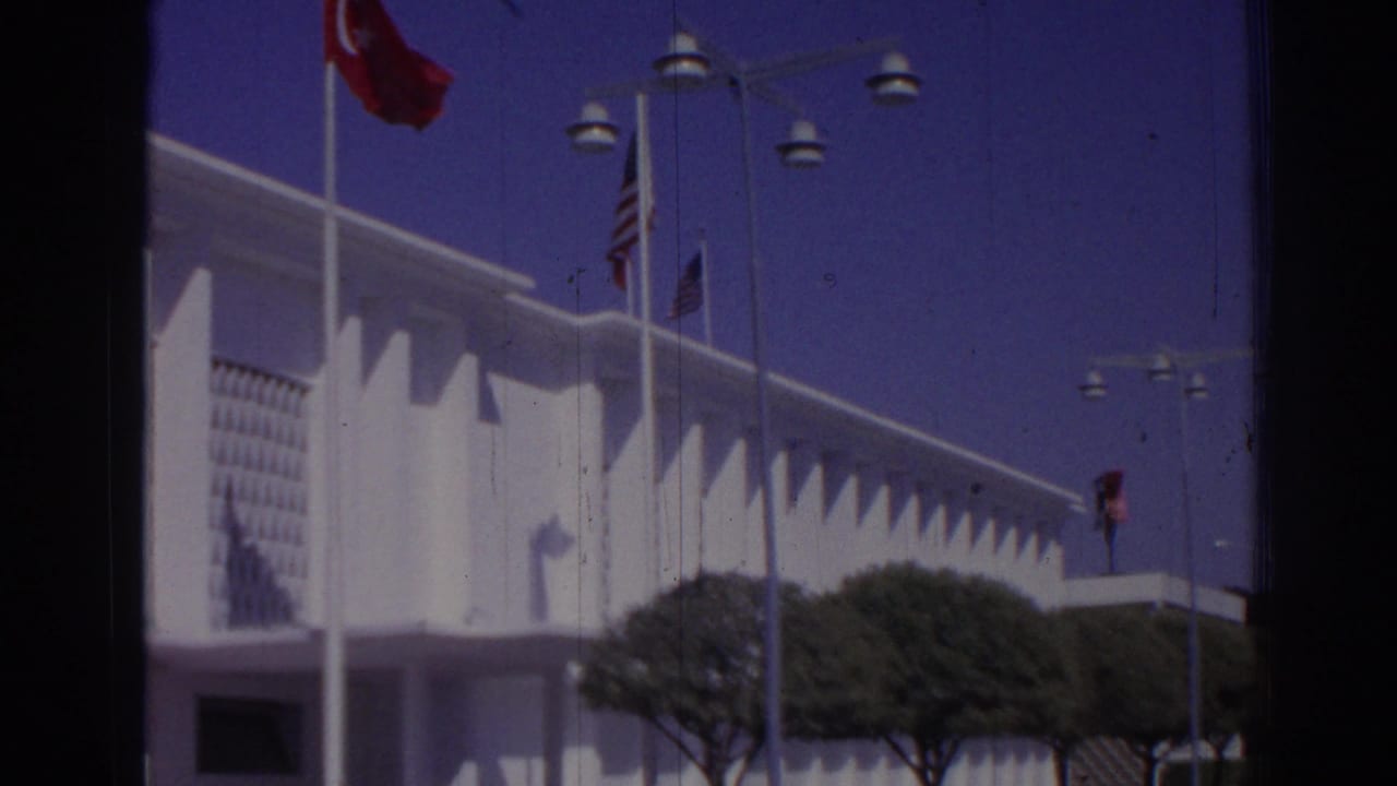 1962 Izmir frame with Turkish and American flags near a white official-looking modern building and trimmed trees.