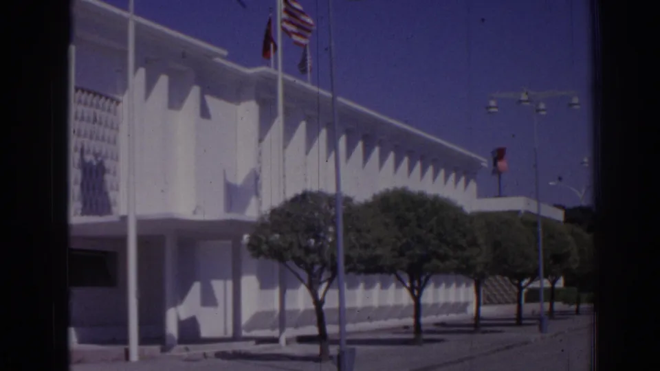 1962 Izmir home-movie view of American and Turkish flags outside a white modernist building with trees and pavement in front.