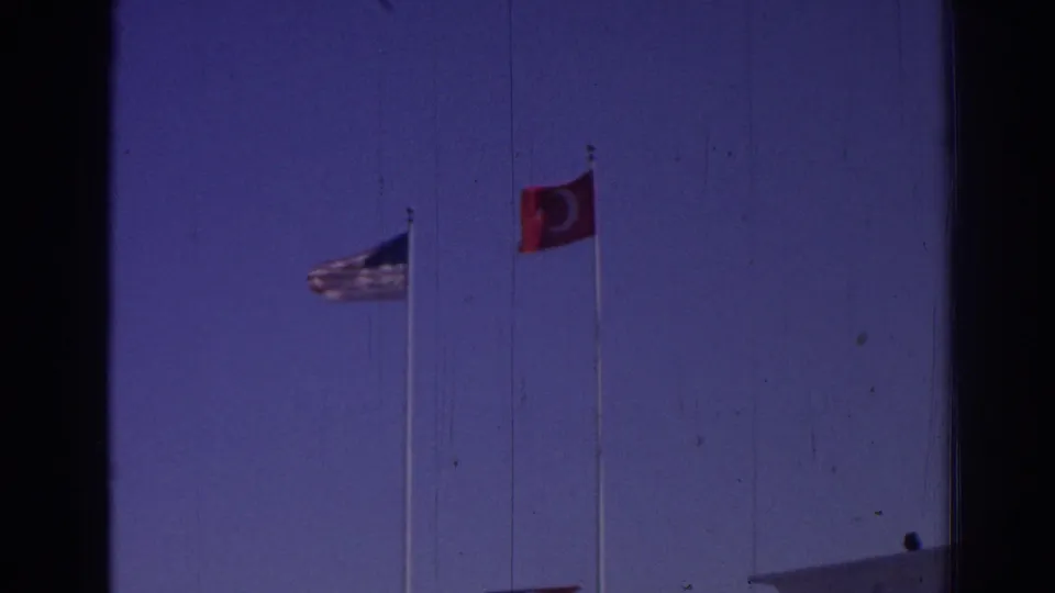 1962 Izmir frame isolating American and Turkish flags on tall poles against a blue sky, with a roof edge at the bottom.