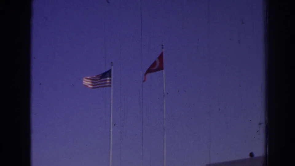 1962 Izmir 8mm frame showing U.S. and Turkish flags above a white building under a blue-purple sky.