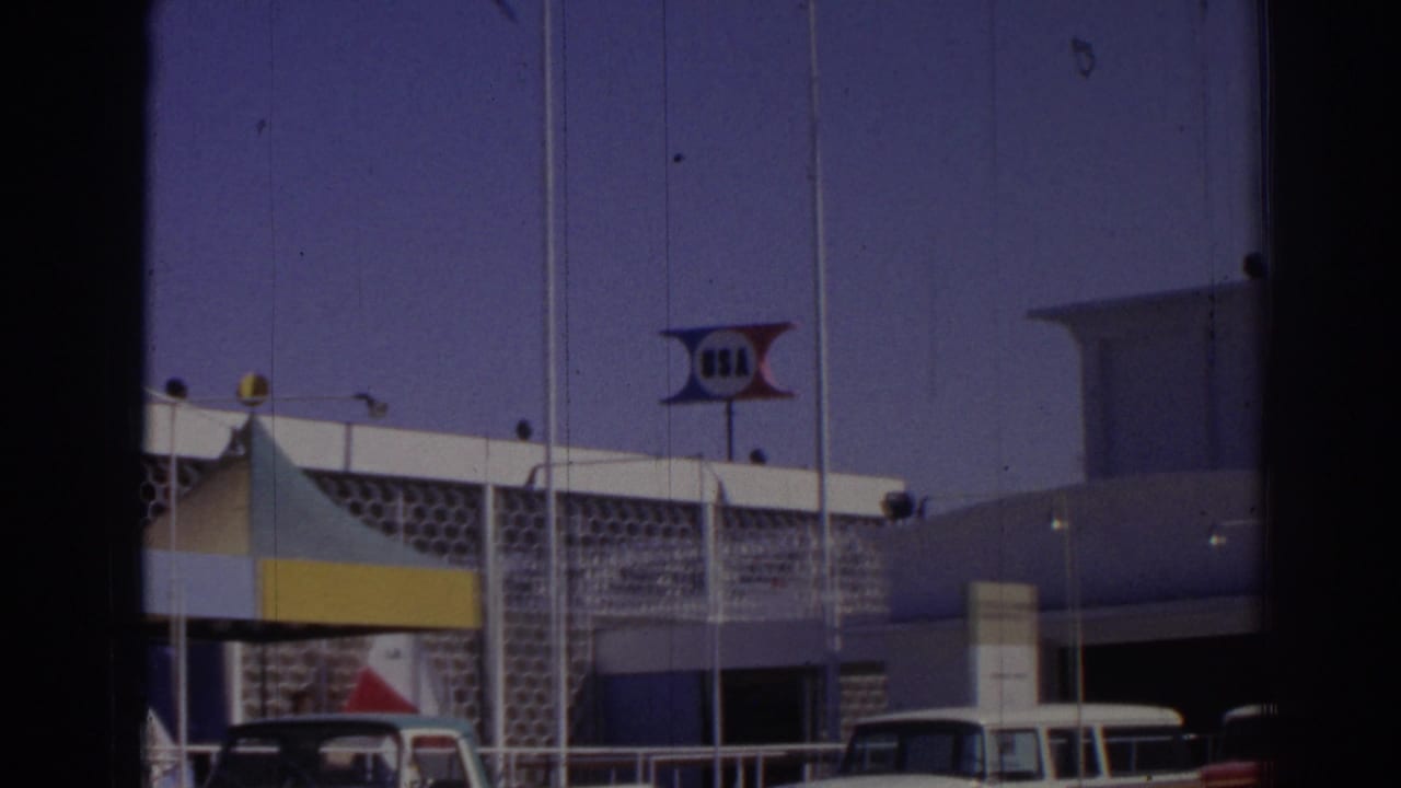 Modernist event building with USA sign, canopy, flagpoles, and parked cars.