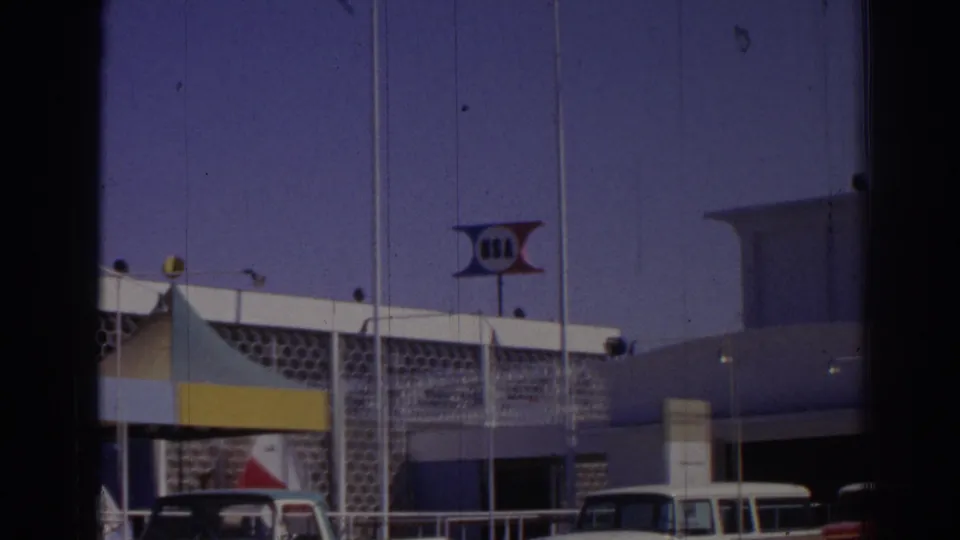 1962 Izmir home-movie frame showing a modernist event building with an apparent USA sign, striped canopy, flagpoles, and parked cars.