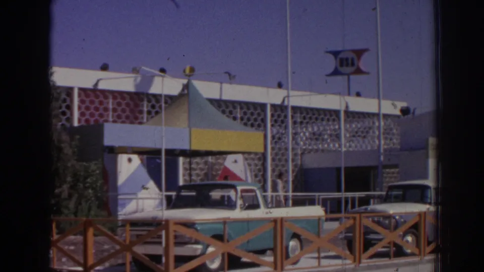 Closer 1962 Izmir view of the apparent USA-signed event building, striped canopy, classic cars, flagpoles, and a wooden foreground rail.