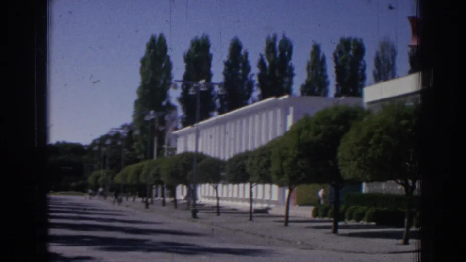 1962 Izmir home-movie frame showing a tree-lined walkway, clipped hedges, a long white institutional building, and small distant people.