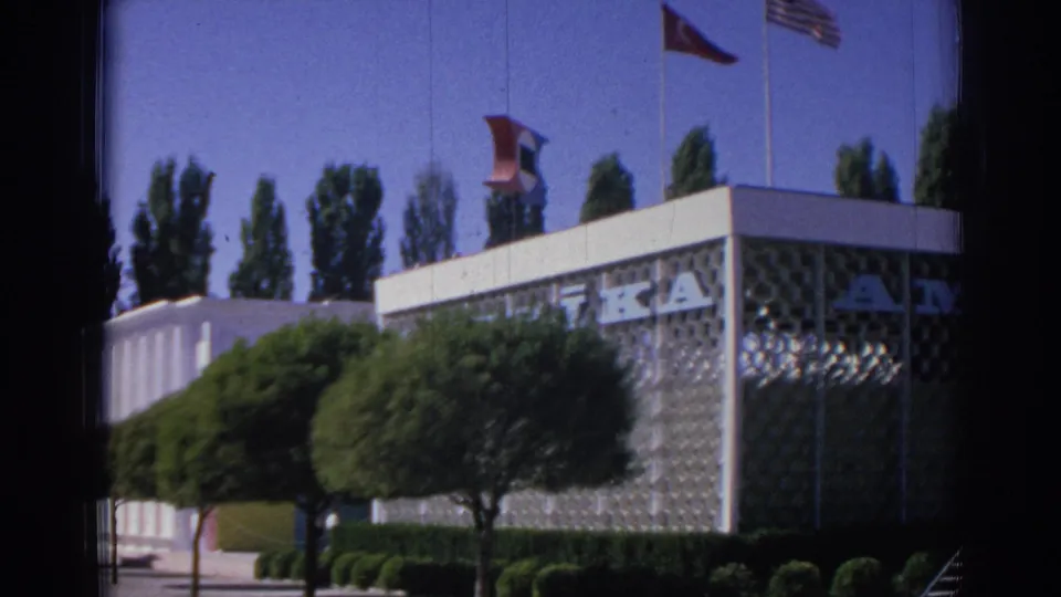 1962 Izmir footage frames a modern corner building with large partial lettering, patterned panels, trees, shrubs, and flags overhead.