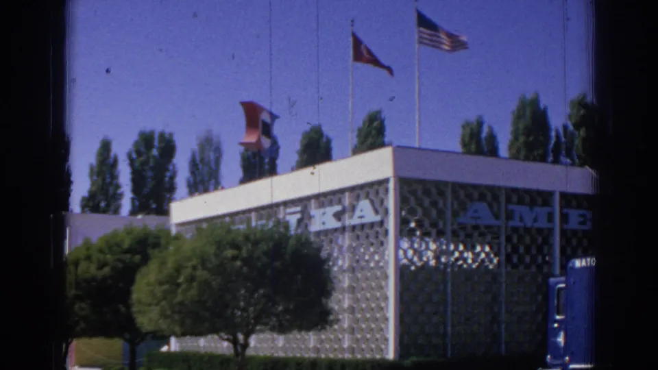 1962 Izmir street view with flags above a modern patterned facade, trimmed greenery, partial lettering, and a vehicle edge at right.