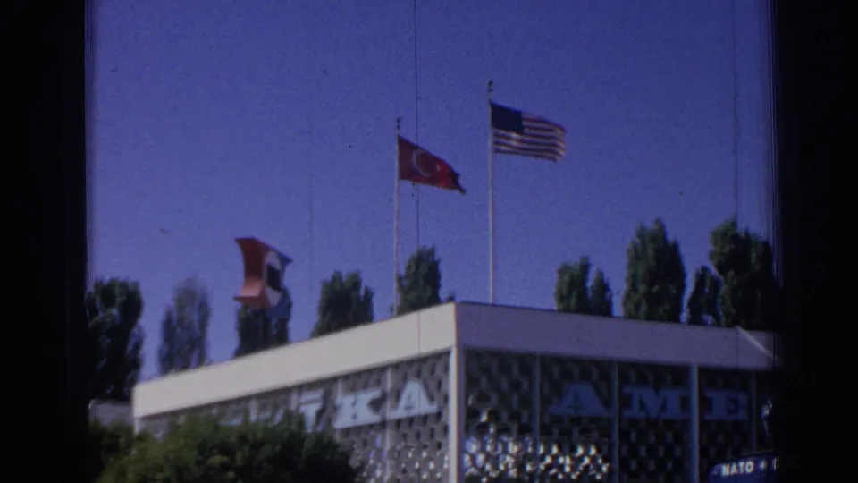 The camera frames flags over the NATO/OTAN facility exterior in Izmir, with the patterned wall and clear sky visible.