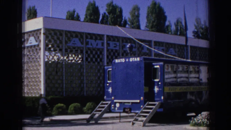 A blue NATO/OTAN-marked transport truck with lowered ramps stands in front of the Izmir facility, with signage and a person nearby.