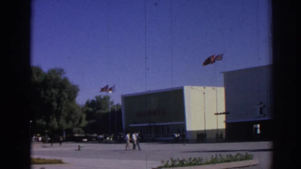 1962 Izmir exterior showing a modern building with a red JAPONTA sign, flags above the entrance area, pedestrians, trees, and parked cars.