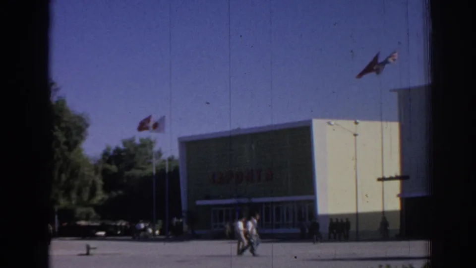 1962 Izmir view of the JAPONTA building facade with flags overhead, people walking across the paved foreground, and trees beside the structure.