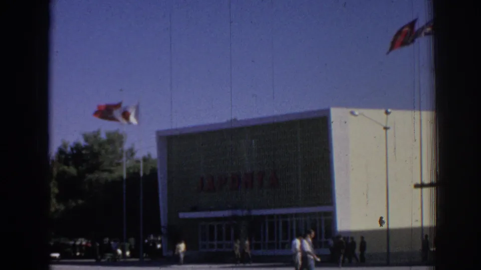 Closer 1962 Izmir frame of the JAPONTA building, showing flags in the sky, glass entry doors, pedestrians near the facade, and dark film borders.