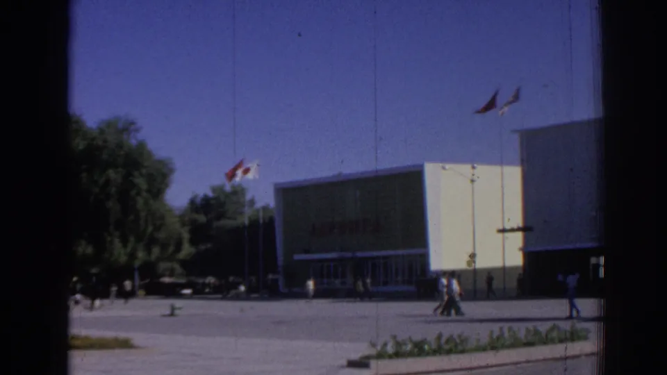1962 Izmir exterior of the JAPONTA building seen across the plaza, with flags above, small groups of pedestrians, trees, and visible 8mm frame edges.
