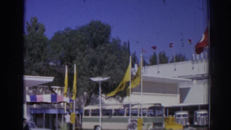 1962 Izmir depot view with a white terminal facade, fluttering flags, parked buses, trees, and a few people at street level.