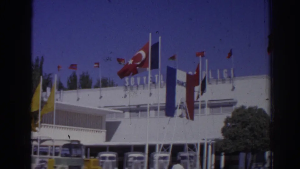 1962 Izmir terminal scene showing roofline signage, multiple flags, parked buses, a tree, and people near the entrance area.