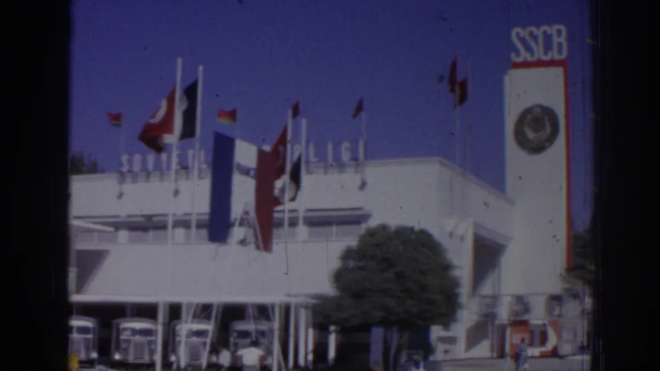 Izmir depot exterior with flags, an SSCB-marked sign panel, parked buses, trees, and pedestrians near the building.