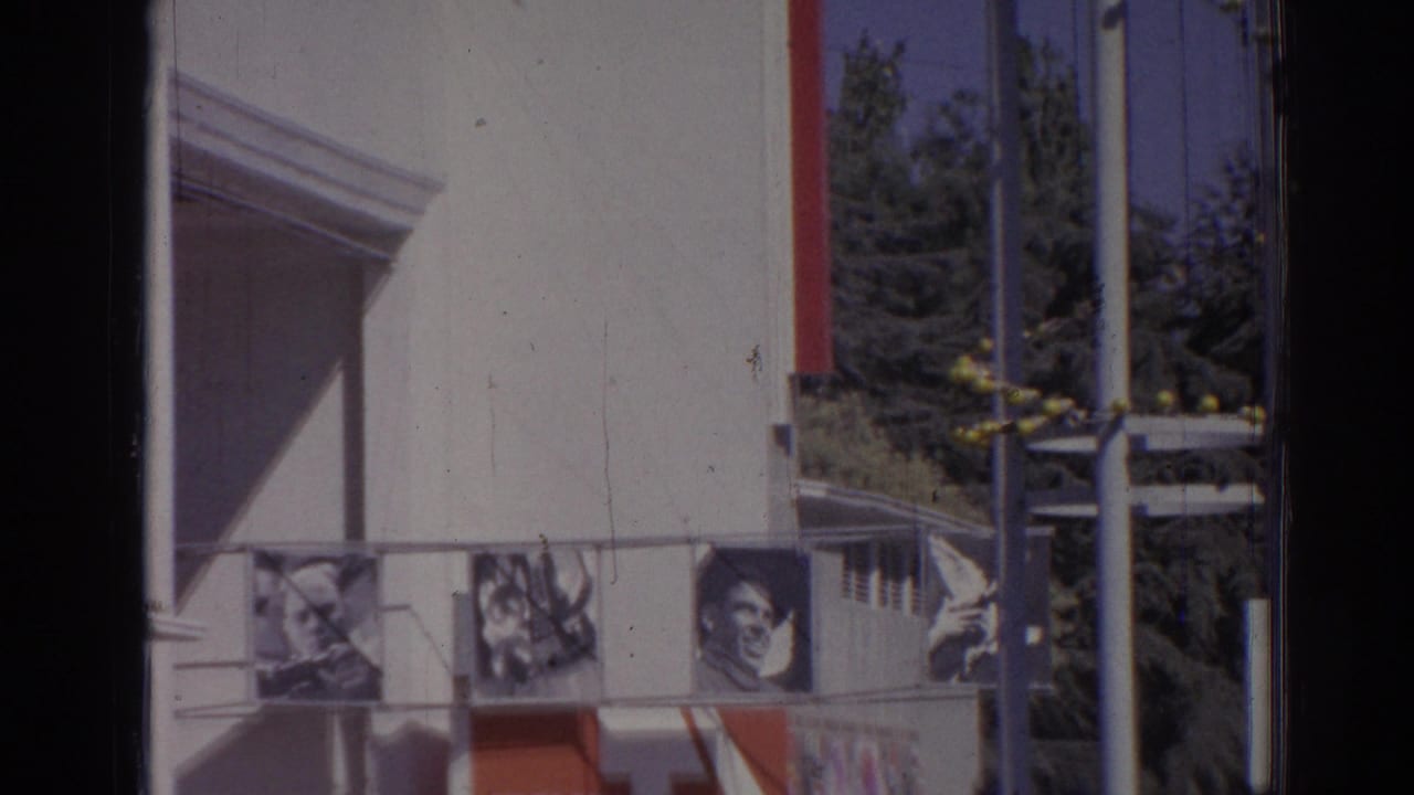 White institutional facade with portrait banners, red trim, blue sky, poles, and trees.