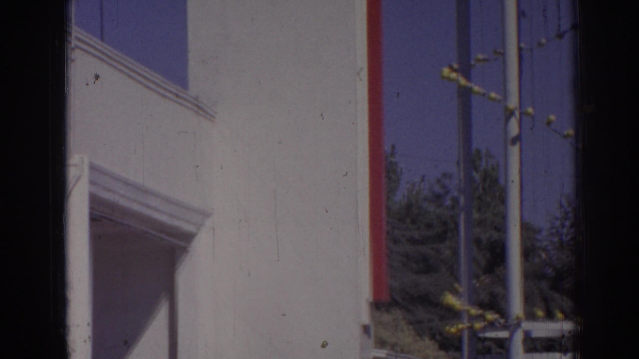 White building wall with red trim, poles, blue sky, trees, and a dark central film edge.