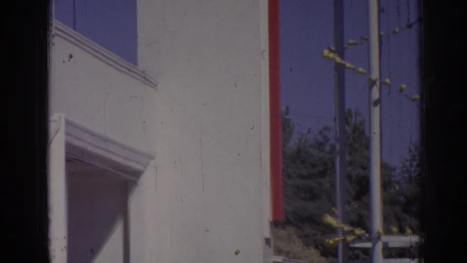 1962 Izmir building facade with a tall white wall, red trim, poles, blue sky, trees, and visible film-frame edge.