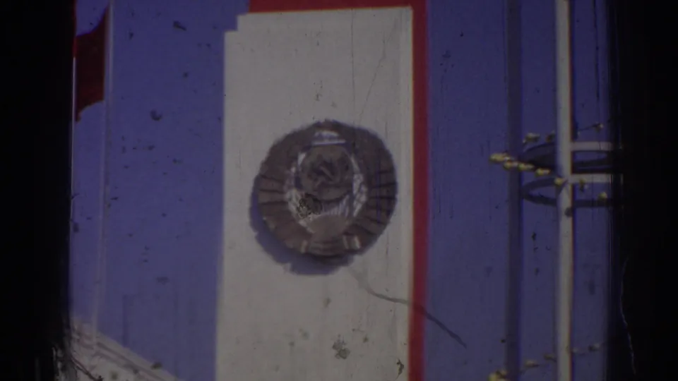 1962 Izmir 8mm frame showing part of a blue-and-white SSCB sign with a circular hammer-and-sickle emblem, red trim, metal poles, and a Turkish flag edge.