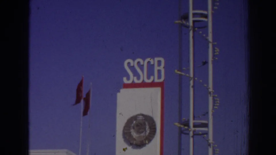 1962 Izmir 8mm frame showing a more distant SSCB sign section with circular emblem, Turkish flag, blue sky, white panel, red stripe, and vertical poles.