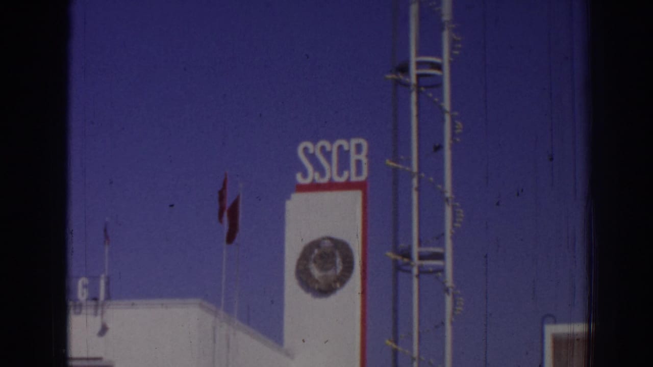 White building tower with SSCB letters, circular emblem, red flags, and blue sky.