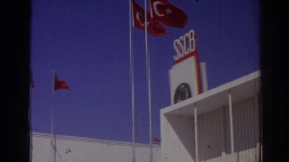 1962 Izmir 8mm frame showing red Turkish flags above a white modern facade with SSCB lettering against a deep blue sky.