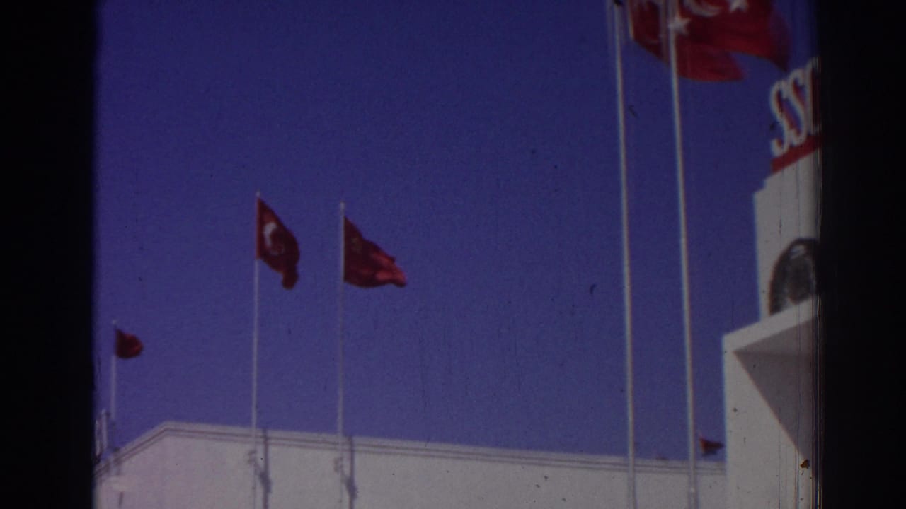 Red flags on tall poles beside part of a white building under blue sky.