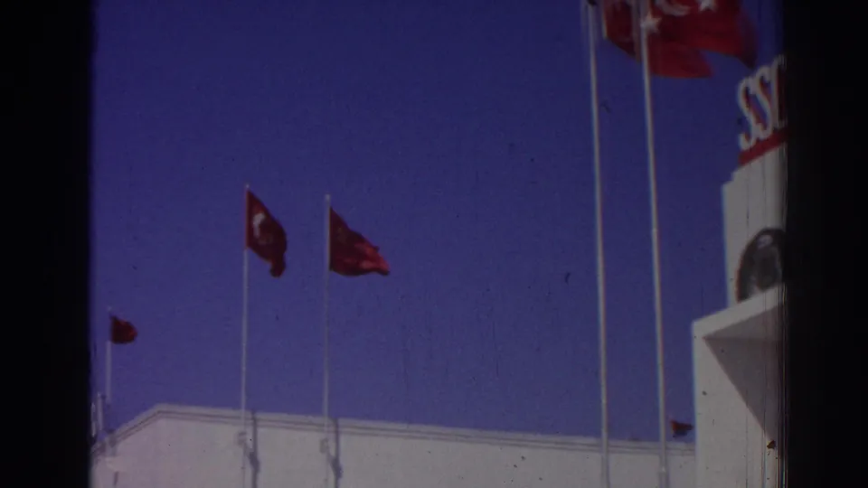 1962 Izmir 8mm frame with red flags on tall poles and the edge of a white building, including a partial SSCB sign at the right.