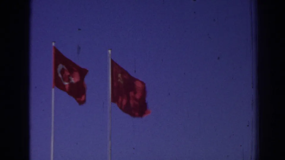 1962 Izmir 8mm frame showing a Turkish flag and a red flag with a yellow emblem waving on tall poles against blue sky.