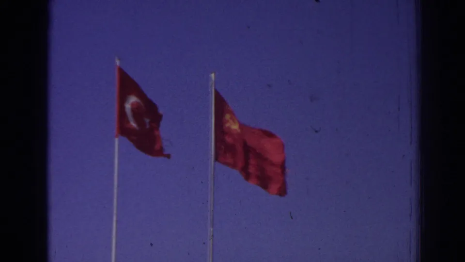 1962 Izmir 8mm frame with a Turkish flag and adjacent red flag bearing a yellow emblem on tall poles against blue sky.