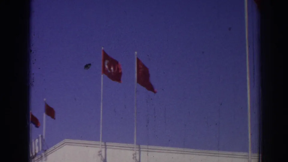 1962 Izmir home-movie frame showing red Turkish flags above a white building roofline under a deep blue sky.