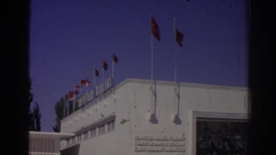 1962 Izmir home-movie view of a white facade, rooftop Turkish flags, and partly legible lettering.