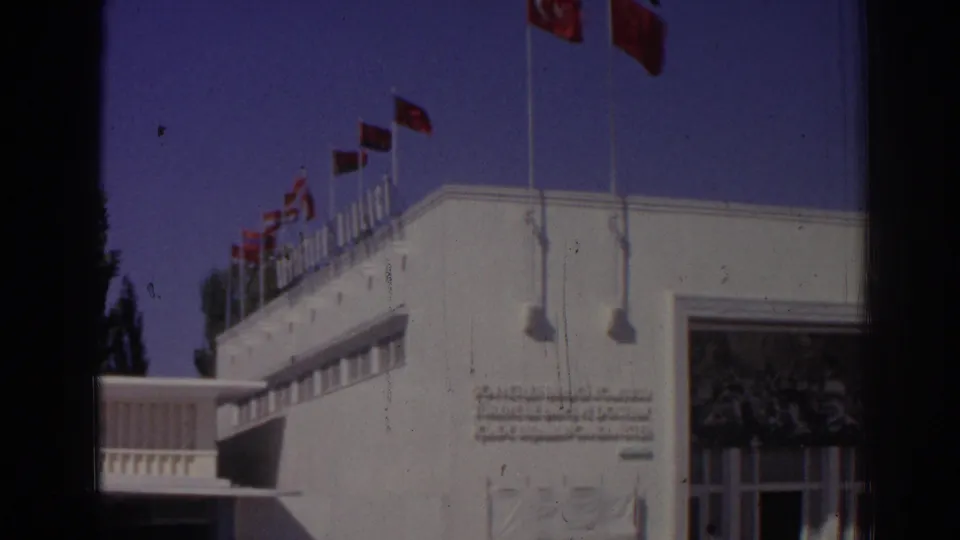 1962 Izmir frame showing a white building corner with rooftop Turkish flags and partial facade inscription.
