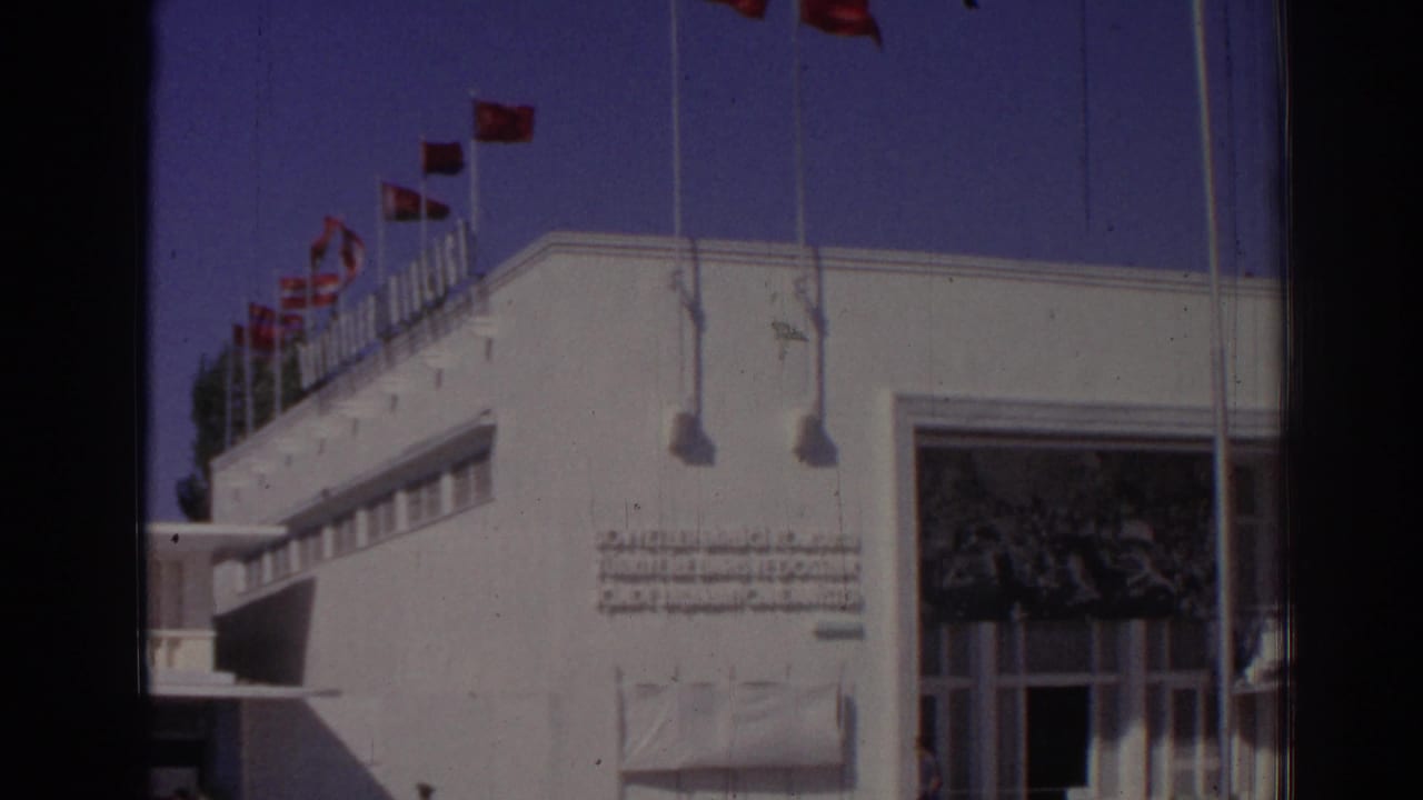 White building facade with rooftop flagpoles and red flags under a blue sky.
