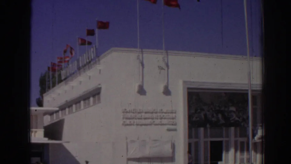 1962 Izmir 8mm frame showing a white modernist building with rooftop red flags, a large entrance, and a Turkish inscription on the facade.