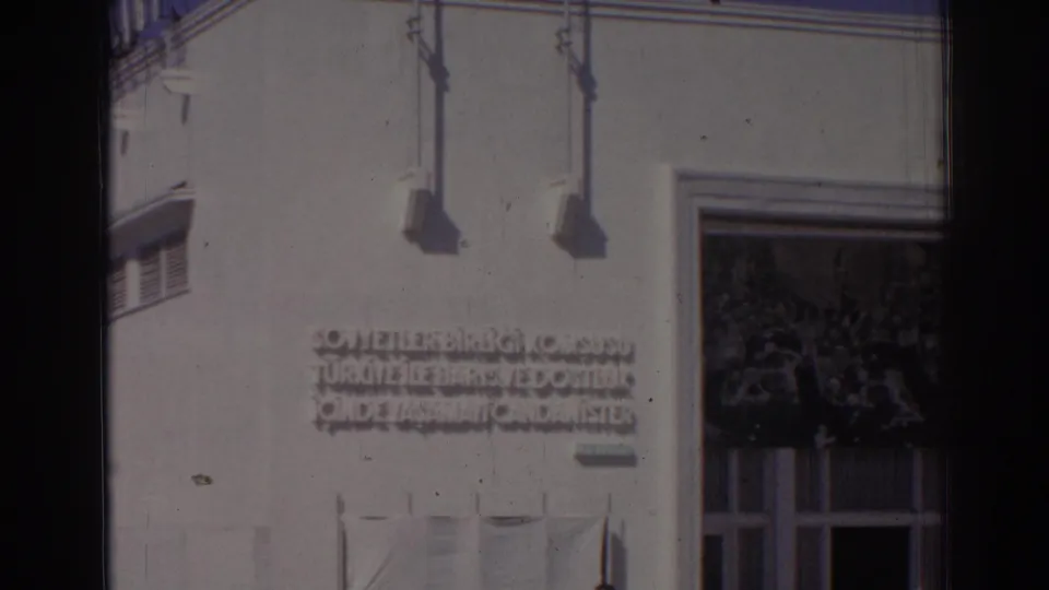 1962 Izmir home-movie frame with the Turkish wall inscription, white facade, flagpole shadows, and the edge of a large entrance.