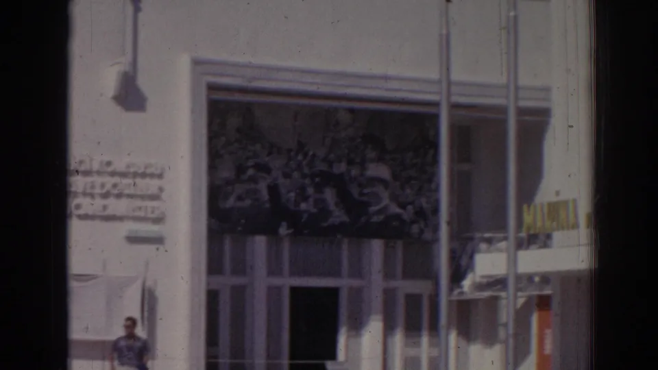 1962 Izmir home-movie frame showing a white facade, crowd mural, wall lettering, poles, and a passerby near the entrance.
