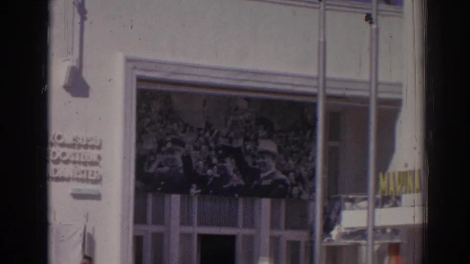 1962 Izmir home-movie frame showing partial Turkish wall text, a crowd mural, facade windows, poles, and a man near the lower edge.